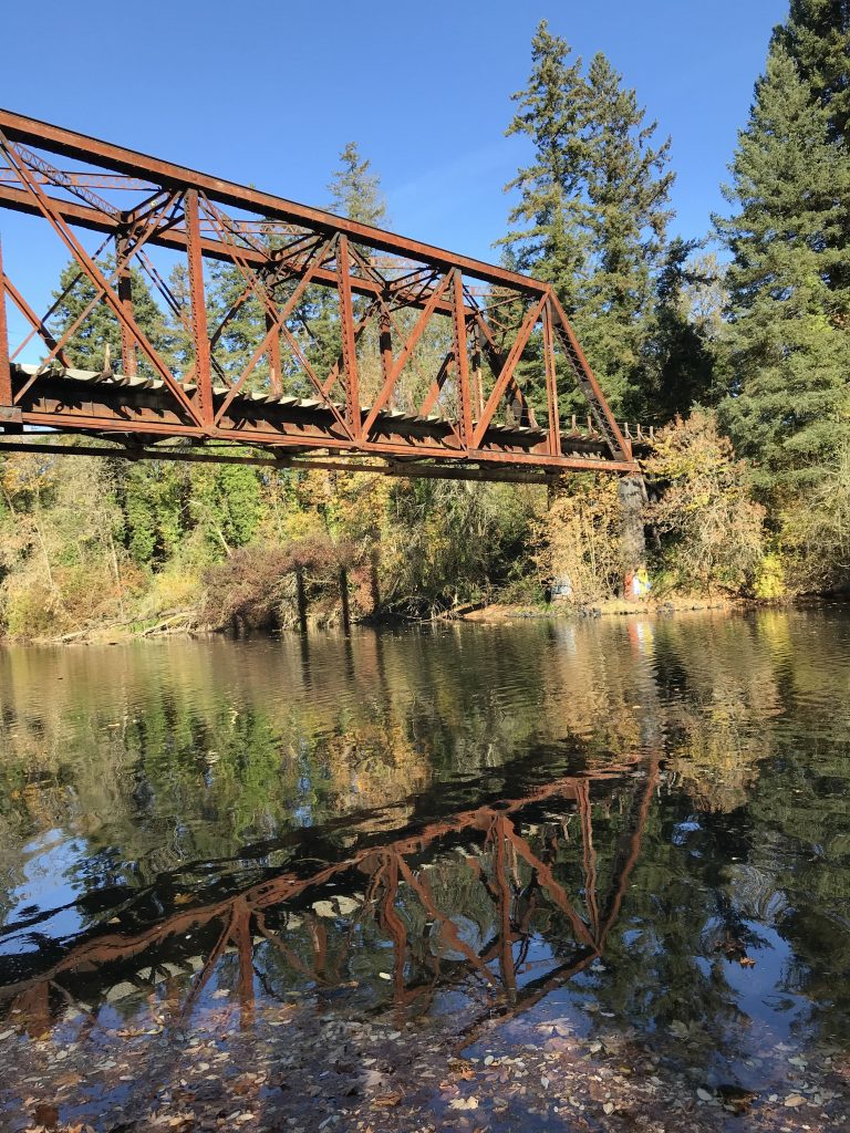 A rusty railroad bridge spanning the Tualatin River. The river is still and dark, the bridge is reflected in the water.