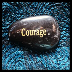 Close up of a shiny, black rock with the word "Courage" written upon it in gold letters. Rock is on blue fabric.